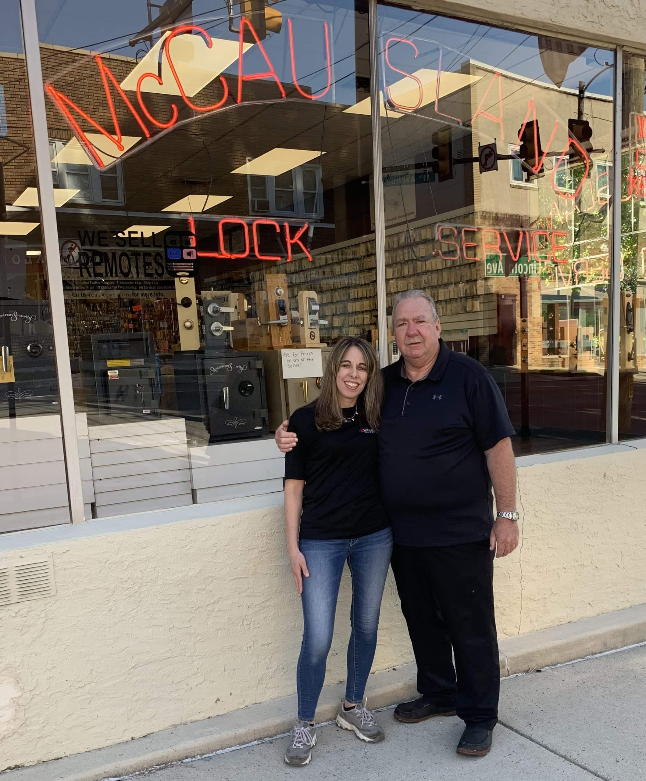 A smiling woman and man stand arm-in-arm on a PA sidewalk in front of a shop with large windows and a neon "LOCK SERVICE" sign, highlighting Locksmith Services Delaware County. Keys and locks are visible inside the store.