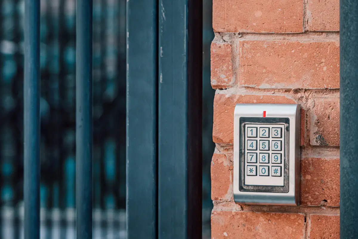 A security keypad is mounted on a brick wall next to a black metal gate. The keypad has numbers 0-9 and extra buttons, and is used for entering an access code to unlock the gate.
