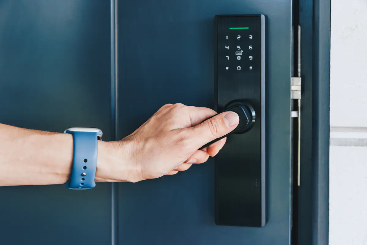 A person wearing a blue smartwatch is unlocking a door using a modern black electronic door lock with a keypad and a fingerprint scanner.