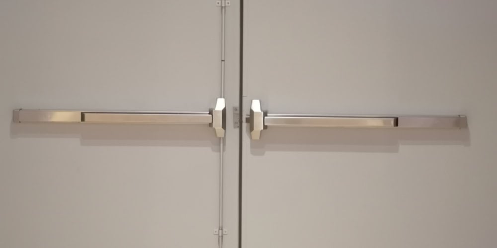 A pair of closed double doors with horizontal push bars, also known as panic bars, typically used for emergency exits. The plain, light-colored doors are commonly serviced by Locksmith Services Delaware County, PA.