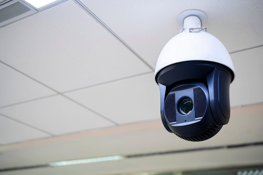 A dome-shaped security camera is mounted on a white ceiling, facing downward in an indoor setting. The camera, ideal for Locksmith Services Delaware County, PA, features a black base and a white cover.