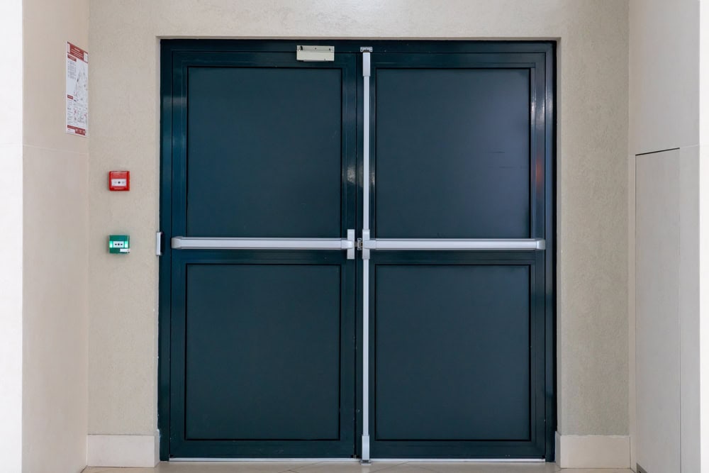 Double emergency exit doors with push bars set in a beige wall. On the left are a red fire alarm and green emergency exit button. A sign is posted above the doors. Locksmith Services Delaware County, PA can assist with secure installation and maintenance.