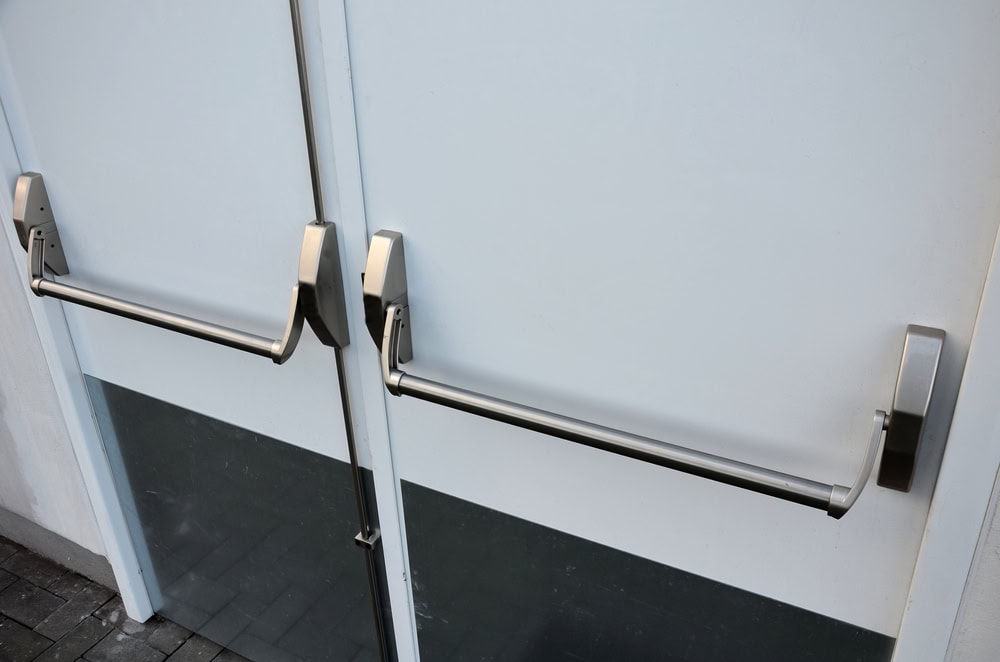 Double white emergency exit doors with horizontal push bars and metallic handles, set against a light background with a dark strip at the bottom—perfect for those seeking secure Locksmith Services in Delaware County, PA.