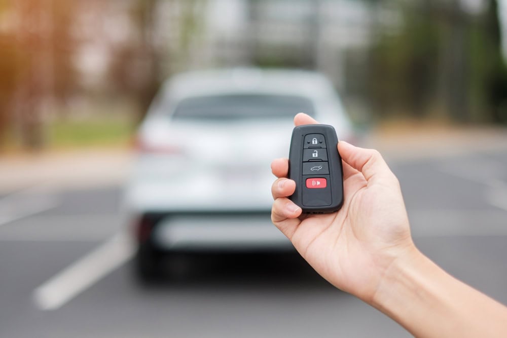 A hand holds a car key remote with buttons for lock, unlock, trunk, and alarm. In the background, a white car sits parked on a blurry outdoor lot in PA—perfect for Locksmith Services Delaware County needs.