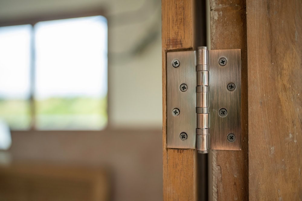Close-up of a metal door hinge attached to a wooden door frame, with a bright, blurred window and room in the background—ideal for showcasing Locksmith Services Delaware County, PA.