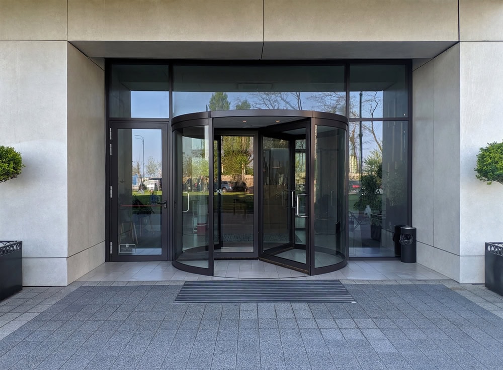 A glass revolving door at the entrance of a modern PA building, flanked by large windows and two round green shrubs in planters on each side. The exterior features light-colored walls and gray paving stones. Locksmith Services Delaware County available nearby.