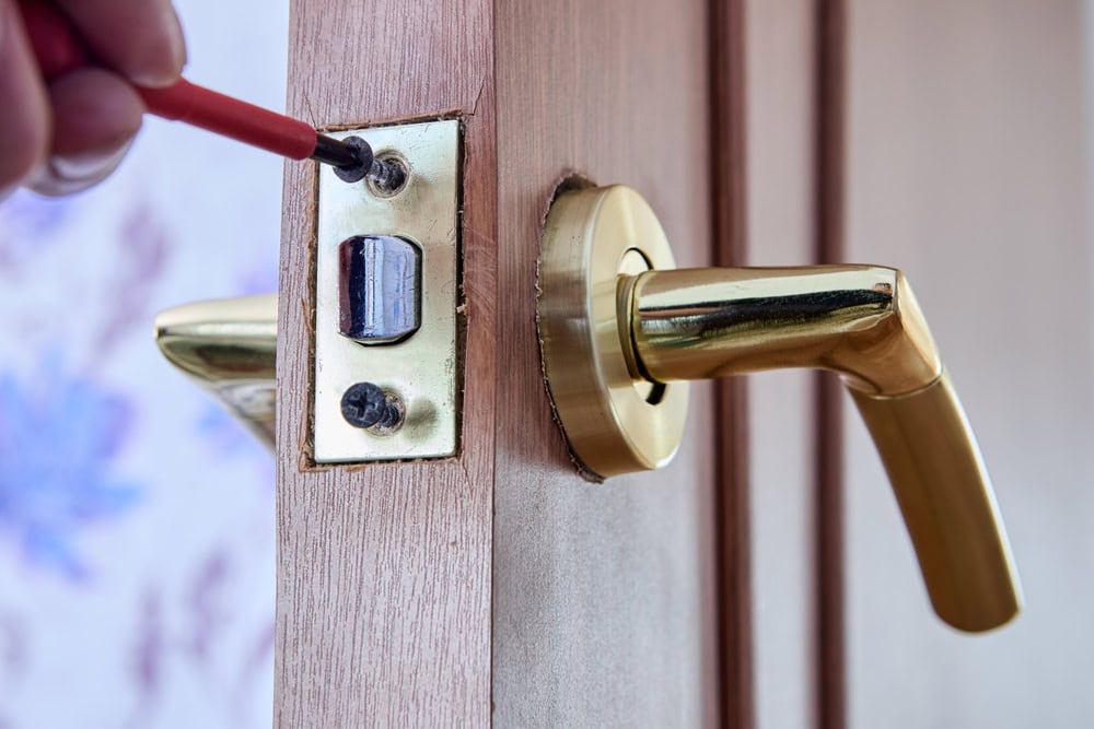 A close-up of a hand using a red screwdriver to tighten a screw on a door latch plate, with a gold-colored door handle attached to a wooden door—professional Locksmith Services Delaware County, PA.
