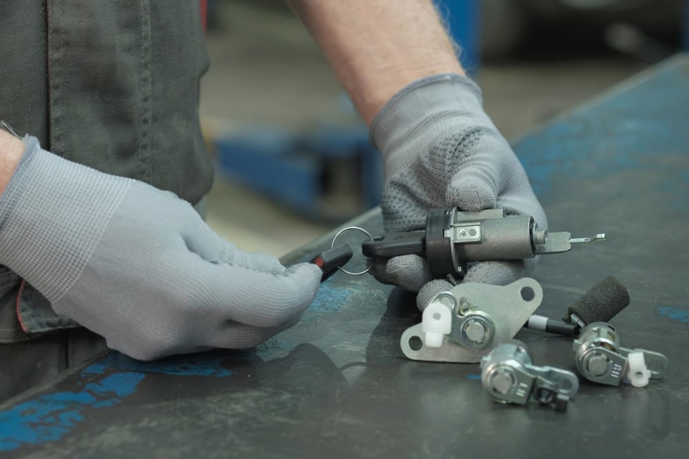 A person wearing gray gloves works with a car ignition lock cylinder and related parts on a metal workbench, using a key and tools for repair or assembly—showcasing professional Locksmith Services Delaware County, PA.