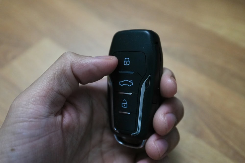 A hand holding a black car key fob with three buttons—lock, unlock, and trunk release—against a wooden background, representing expert Locksmith Services Delaware County, PA.