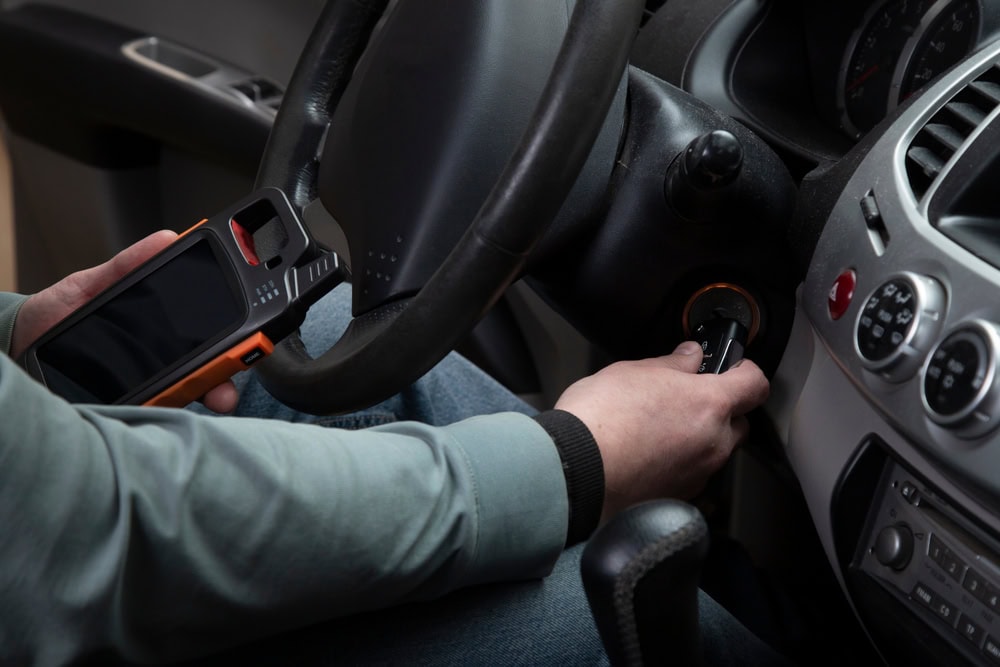 A person sitting in a car uses one hand to insert a key into the ignition, while holding an electronic device with a screen and buttons—possibly related to Locksmith Services Delaware County, PA—in the other hand near the steering wheel and dashboard.