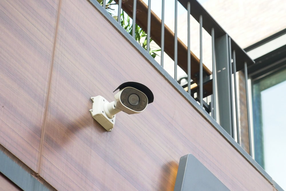 A security camera mounted on a wooden exterior wall, angled slightly downward—ideal for enhancing safety alongside Locksmith Services Delaware County, PA. Black metal railings and plants are visible above as sunlight shines on the building.