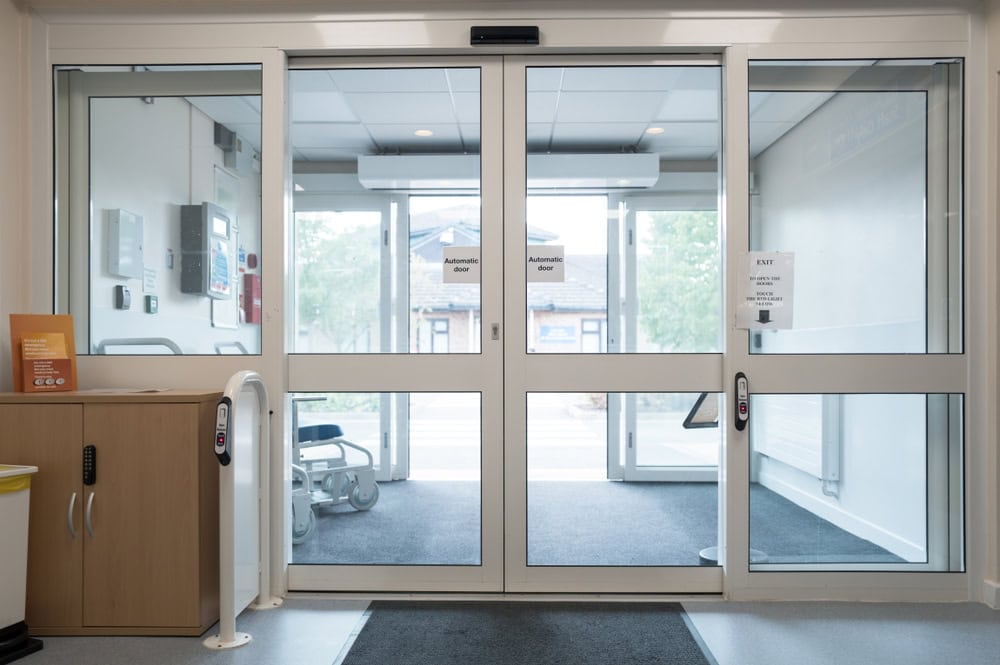 Automatic glass double doors at the entrance of a modern building, with "Automatic Door" signs, a wooden cabinet on the left, and medical equipment in the background—ideal for those seeking locksmith services in Delaware County, PA.