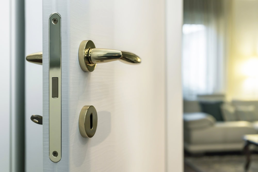 A close-up of a slightly open white door with a shiny gold handle and lock, revealing part of a softly-lit PA living room with a sofa and lamp in the background, perfect for showcasing Locksmith Services Delaware County.
