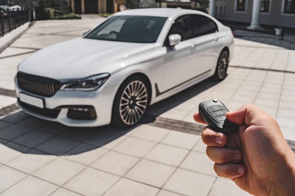A person holds a car key remote, pointing it towards a parked white luxury sedan on a tiled driveway in front of a building, showcasing Locksmith Services Delaware County, PA.
