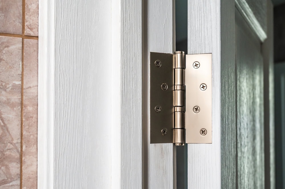 A close-up of a metallic door hinge attached to a white wooden door and frame, adjacent to a tiled wall. The hinge is securely fastened with screws—showcasing expert installation typical of Locksmith Services Delaware County, PA.