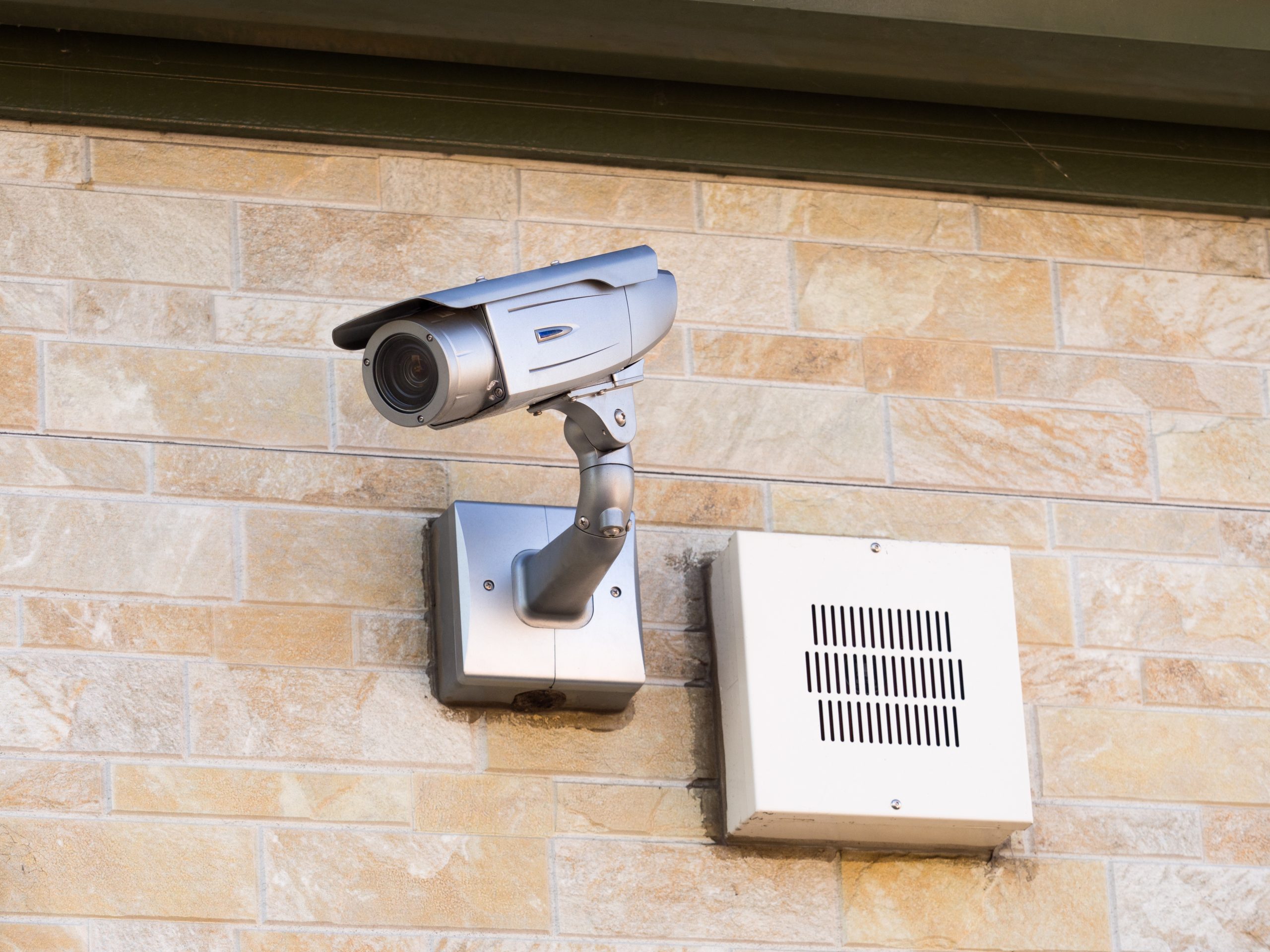 A silver security camera is mounted on a beige brick wall next to a white rectangular speaker or vent box, providing vigilant surveillance—an essential feature for businesses using Locksmith Services Delaware County, PA.