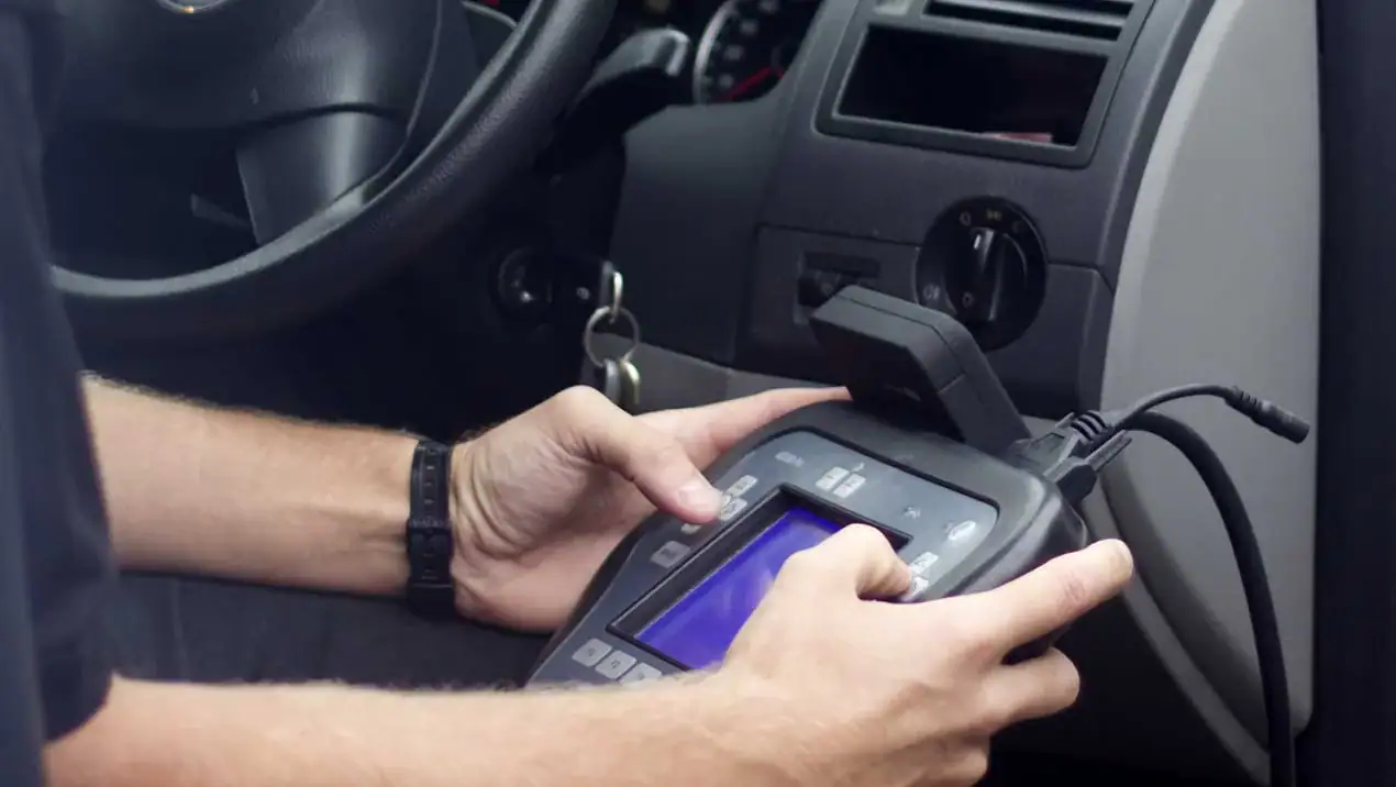 A person sits in a car using a diagnostic tool connected to the vehicle’s dashboard, likely checking or resetting system codes near the steering wheel and ignition.