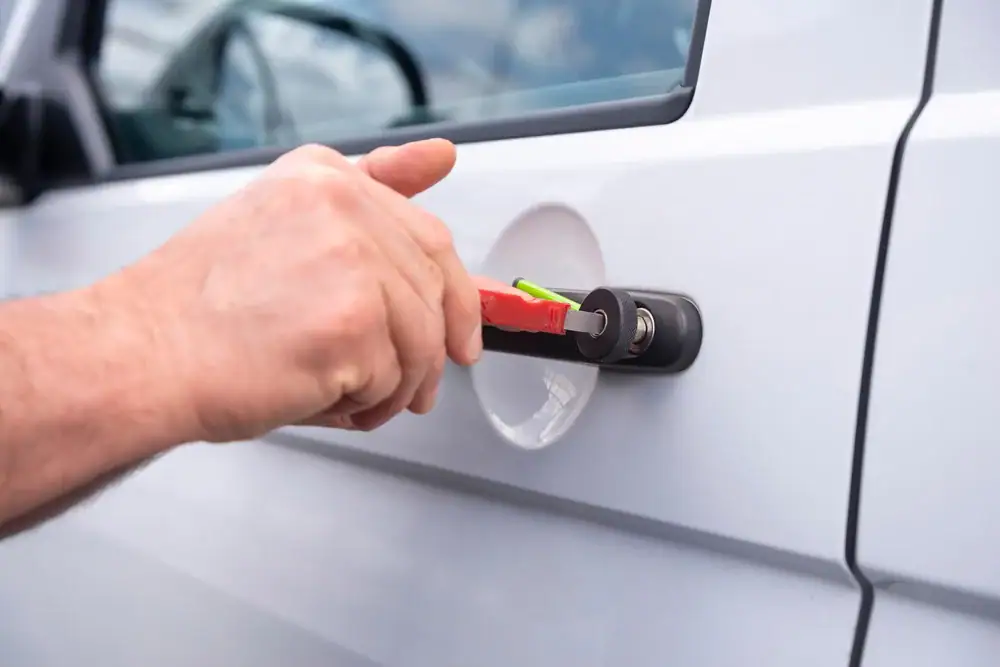 A person&rsquo;s hand is using a red tool to pick or tamper with the lock on a white car door.
