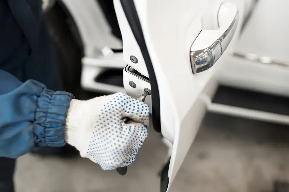 A person wearing white gloves and a blue sleeve uses a screwdriver to adjust or repair the hinge of a white car door.