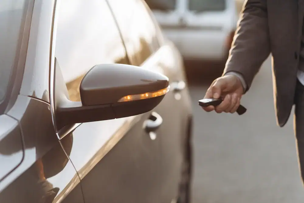 A person wearing a suit holds a key fob while standing next to a parked car, with the car's side mirror and indicator light visible in the foreground.