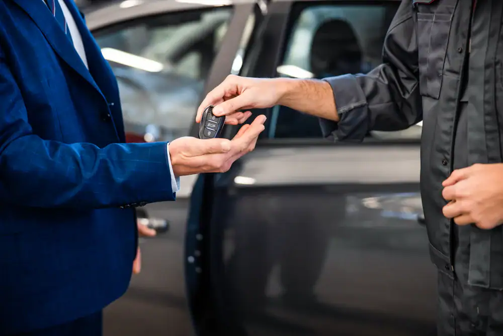 A person in a suit receives a car key from another person in a jumpsuit, standing beside a parked car with an open door.