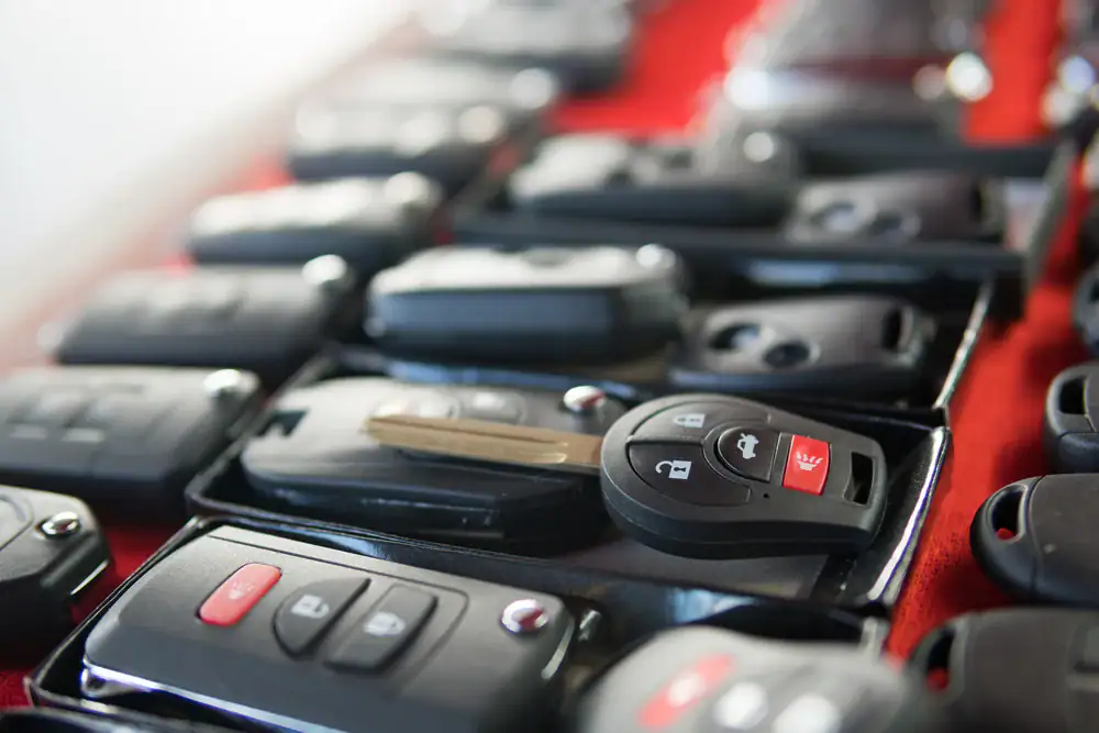 A close-up of various car key fobs and remote controls arranged in rows on a red surface. One key with a metal blade is prominent among the mostly black key fobs.
