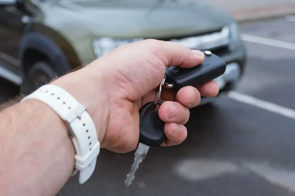 A person&rsquo;s hand holding a car key and remote, pointing it toward a parked, blurred green SUV in an outdoor parking lot. The person is wearing a white wristwatch.