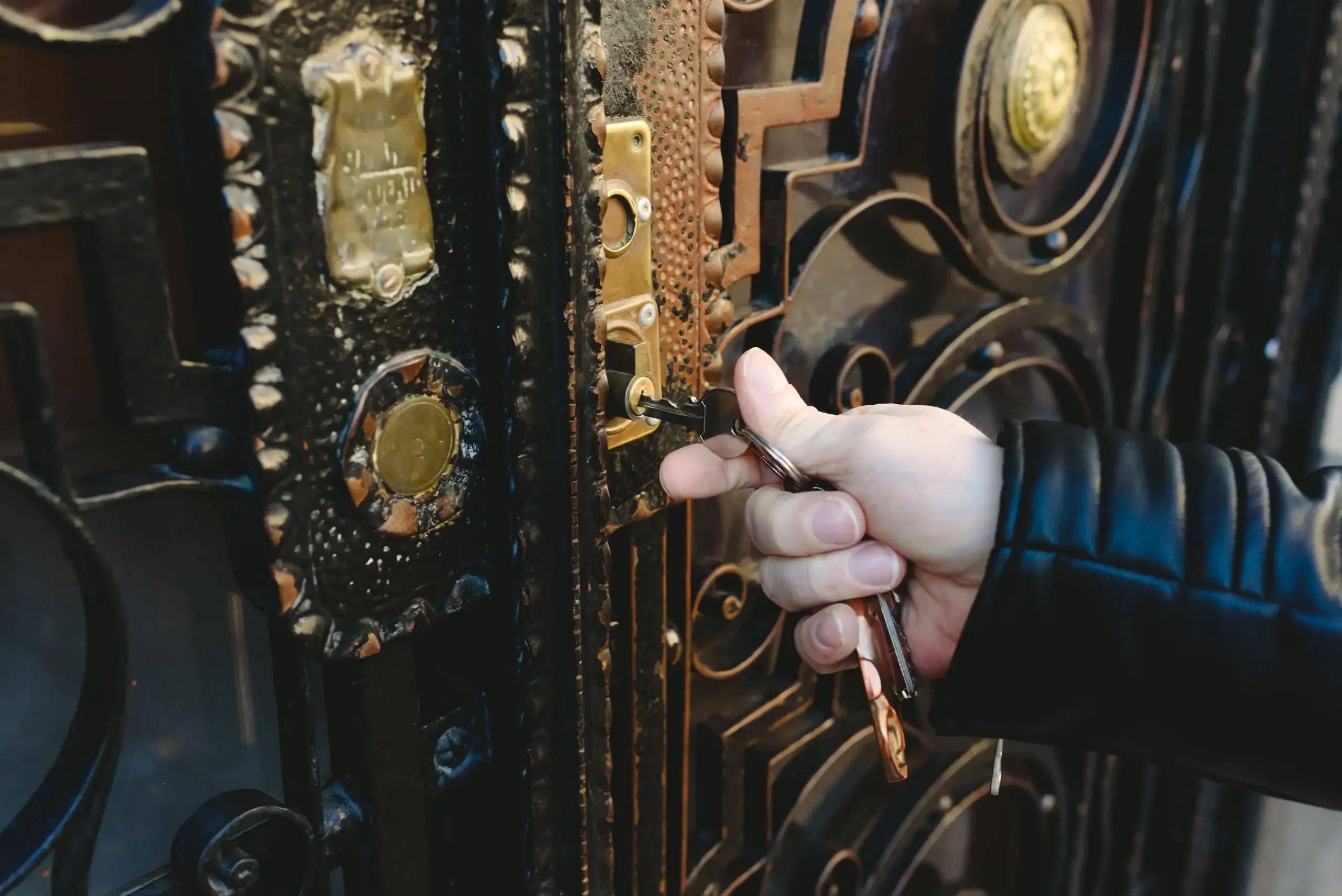A person wearing a black leather jacket is inserting a key into the lock of an ornate, decorative metal door with brass details.