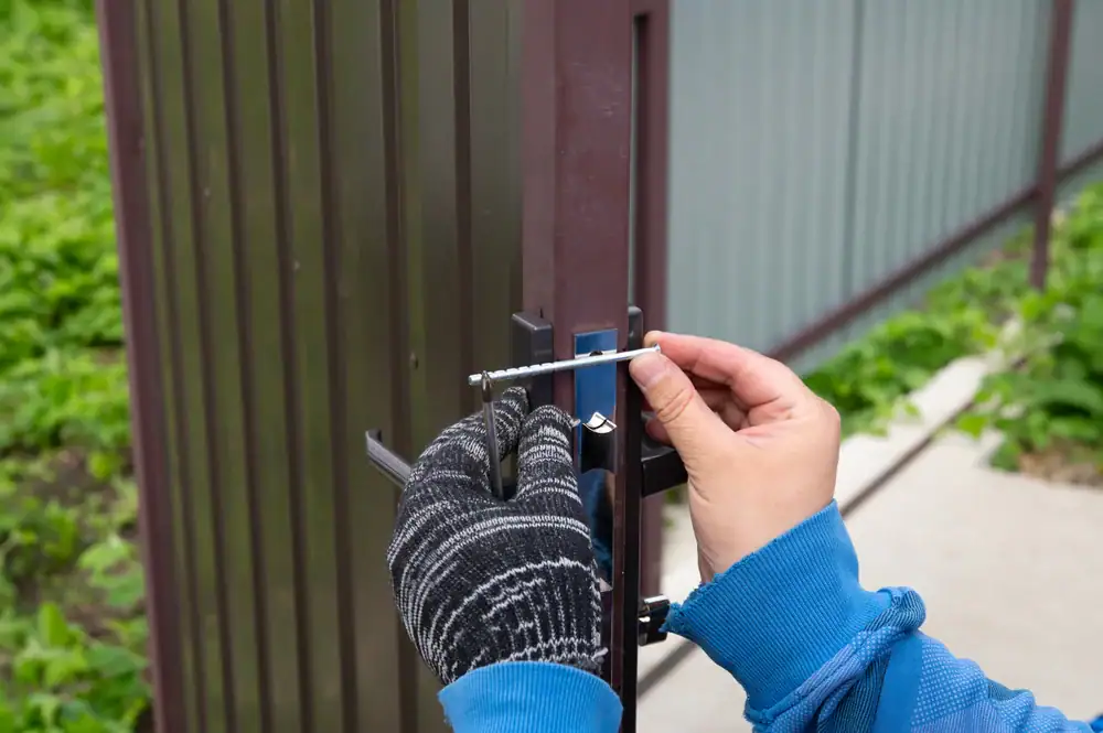 A person wearing a blue sleeve and a black-and-white glove is installing or adjusting a metal latch on a brown metal gate outdoors, using a screwdriver.