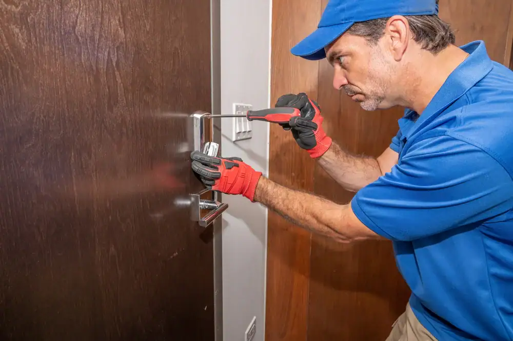 A locksmith wearing red gloves, a blue shirt, and a blue cap is fixing a door lock with a screwdriver and pliers. The door is brown and partially open.
