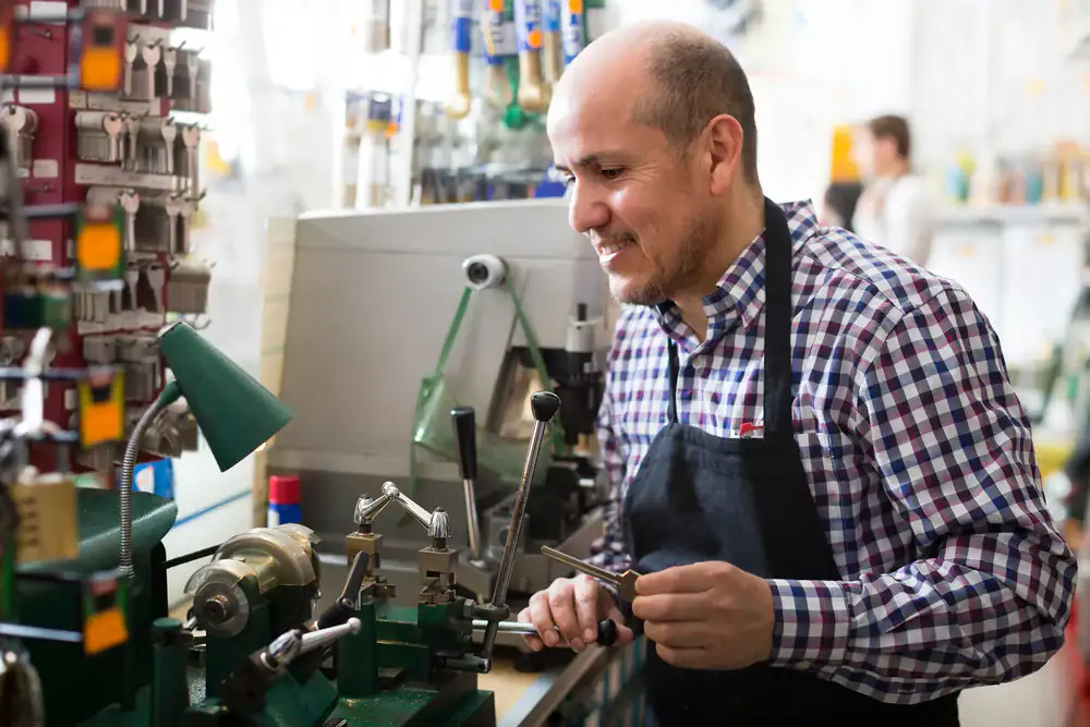 A smiling locksmith in a checkered shirt and apron works at a key cutting machine in a shop, surrounded by hanging keys and various locksmith tools.