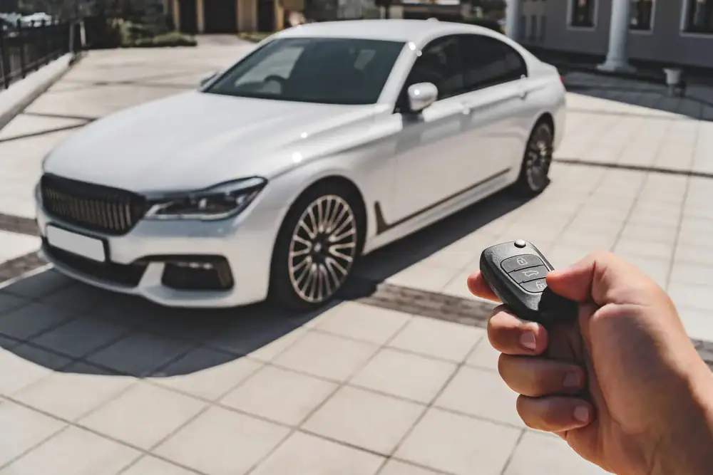 A person holds a car key fob, pointing it toward a white luxury sedan parked on a paved driveway in front of modern buildings.
