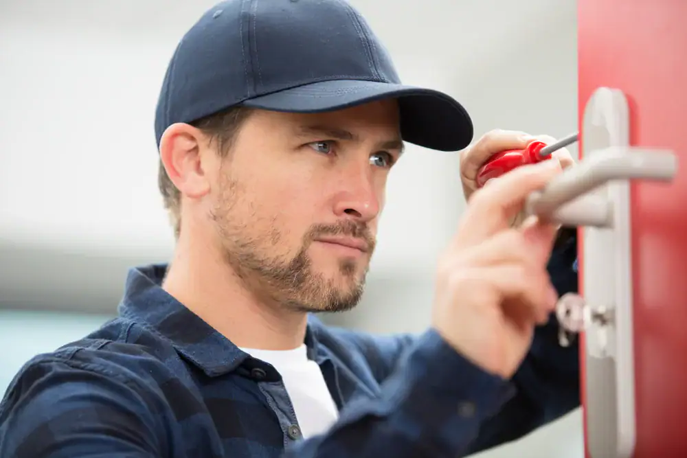 A man wearing a navy cap and plaid shirt uses a screwdriver to fix or install a door handle on a red door.