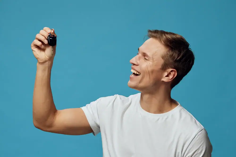 A smiling man in a white t-shirt holds up a car key, looking at it with excitement, against a plain blue background.