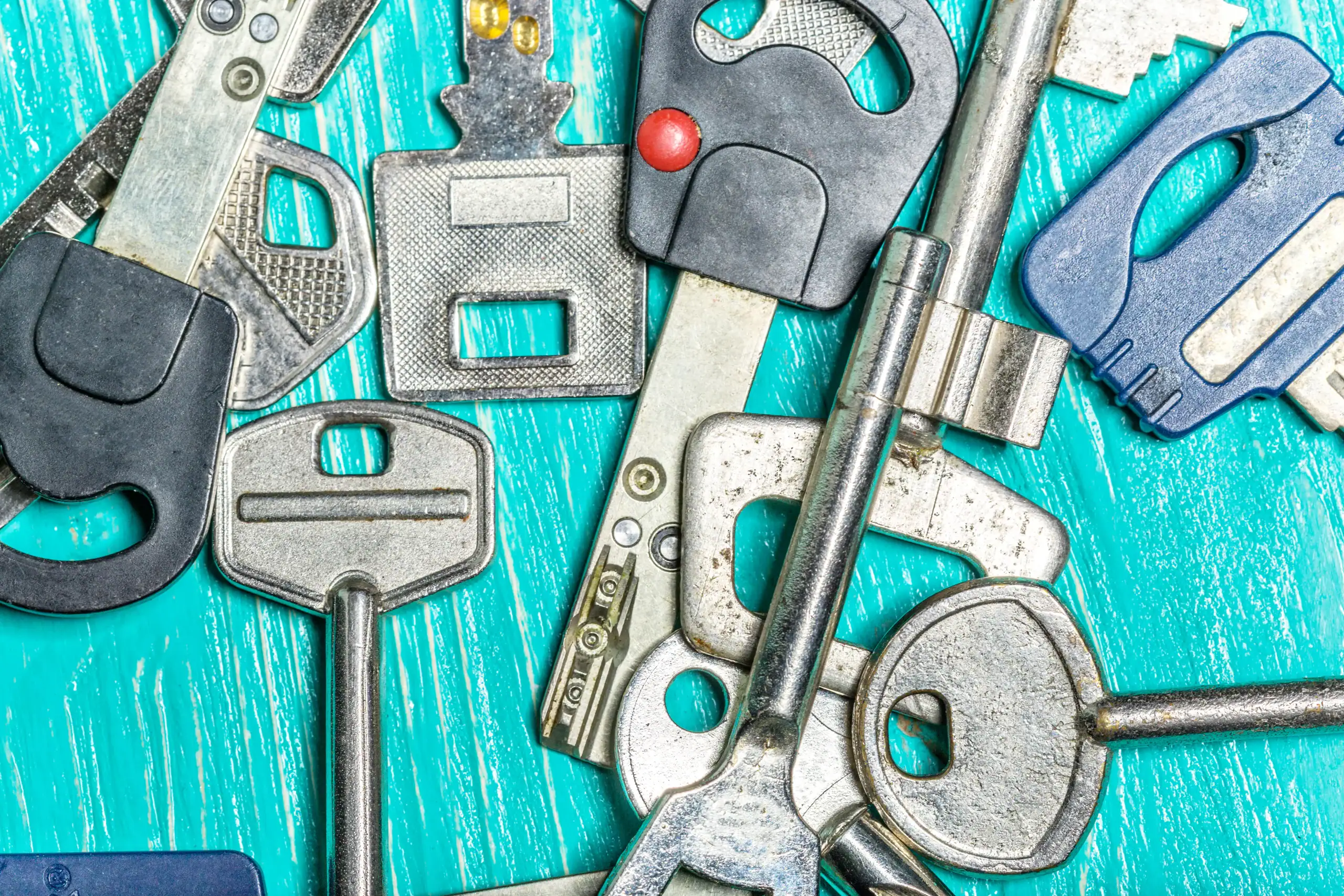 A variety of metal keys, including car and house keys, are scattered on a bright turquoise wooden surface. The keys differ in shapes, sizes, and designs.