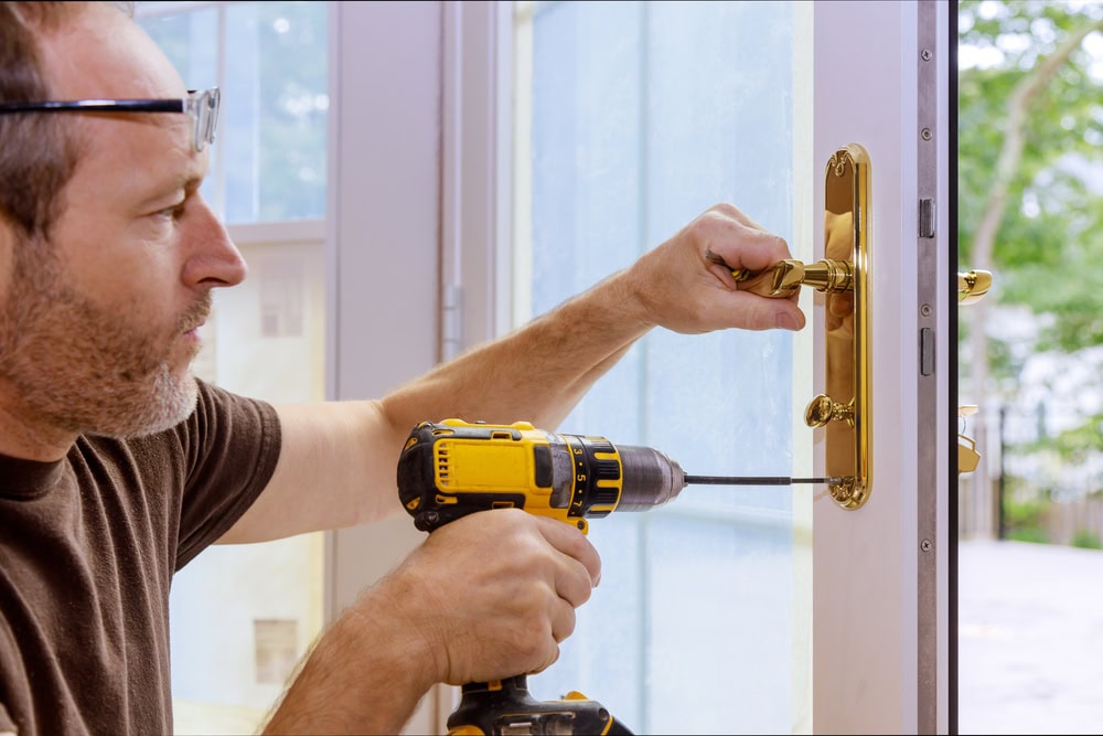 A person uses a yellow cordless drill to install or tighten a gold door handle and lock on a glass door, focusing intently on their work—showcasing expert Locksmith Services in Delaware County, PA.
