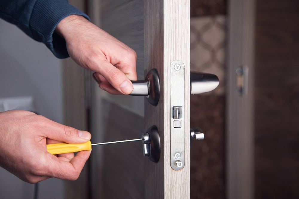 A person uses a yellow-handled tool to pick the lock of a partially open wooden door in PA, while holding the door handle with the other hand—ideal imagery for Locksmith Services Delaware County.