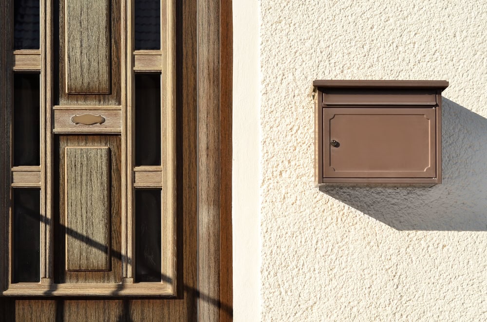 A brown mailbox is mounted on a textured beige wall next to a wooden front door with decorative panels. Sunlight casts shadows, highlighting the entry—a welcoming sight for those seeking Locksmith Services in Delaware County, PA.