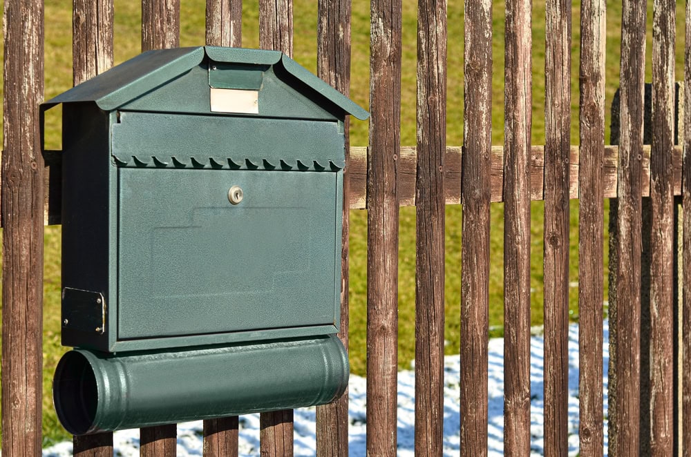 A green metal mailbox with a cylinder-shaped newspaper holder is mounted on a wooden fence with vertical slats. Grass and a small patch of snow are visible—ideal for residents seeking Locksmith Services Delaware County, PA.