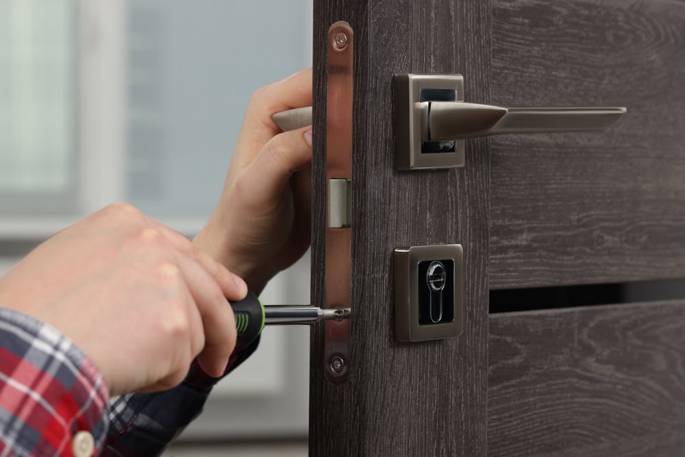 A person in a plaid shirt uses a screwdriver to install or repair the lock mechanism on a dark wooden door, showcasing professional locksmith services in Delaware County, PA. The door is partially open, with sleek silver-colored hardware.