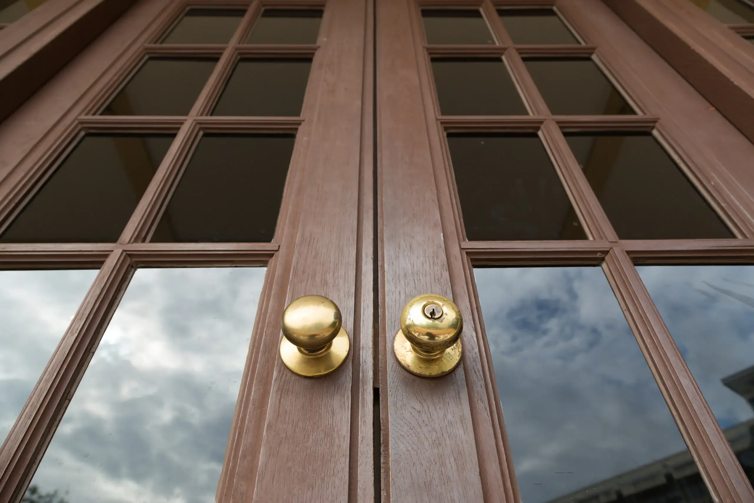 Close-up view of two brass door knobs on wooden double doors with glass panels reflecting a cloudy sky.
